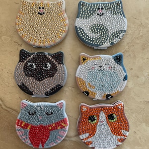 May include: Six compact mirrors with different cat designs. Each mirror is decorated with rhinestones and features a different cat face. The cats are a light brown tabby, a gray tabby, a black and white cat, a calico cat, a gray cat with a red scarf, and an orange tabby.