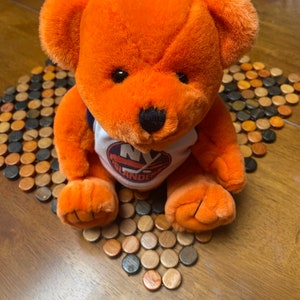 May include: Orange plush teddy bear wearing a white shirt with the New York Islanders logo. The bear is sitting on a wooden tile floor.