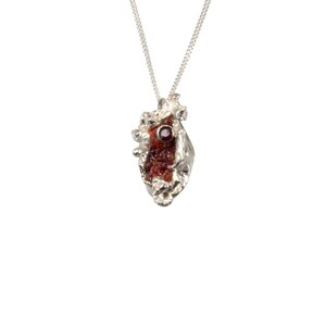 Puede incluir: Un collar de plata con un colgante en forma de corazón texturizado en plata con una piedra preciosa roja.