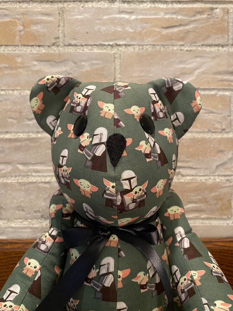 Puede incluir: Un oso de peluche verde con una cinta negra alrededor del cuello. El oso est&aacute; cubierto de un patr&oacute;n de peque&ntilde;as im&aacute;genes repetidas de un personaje de la franquicia Star Wars, The Mandalorian, sosteniendo una peque&ntilde;a criatura alien&iacute;gena verde, conocida como The Child o Baby Yoda.