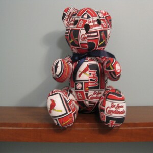 May include: A red, white and blue teddy bear with a St. Louis Cardinals pattern. The bear is sitting on a wooden shelf.
