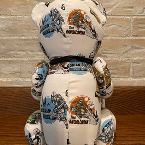 Op de afbeelding: Witte pluche teddybeer met een zwart lint om zijn nek. De beer is bedekt met een stoffen patroon met het Mandalorian-personage uit de Star Wars-franchise. Het stoffen patroon bevat de woorden "The Mandalorian" en "The Mandalorian" die meerdere keren worden herhaald.