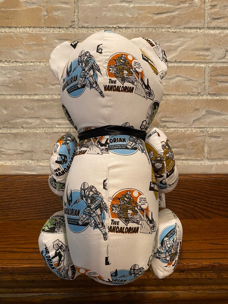 Op de afbeelding: Witte pluche teddybeer met een zwart lint om zijn nek. De beer is bedekt met een stoffen patroon met het Mandalorian-personage uit de Star Wars-franchise. Het stoffen patroon bevat de woorden "The Mandalorian" en "The Mandalorian" die meerdere keren worden herhaald.