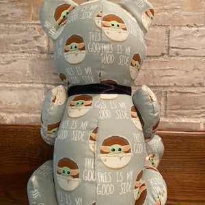 K&ouml;nnte beinhalten: Ein Pl&uuml;schteddyb&auml;r mit einem hellblauen Stoff, der ein sich wiederholendes Muster eines gr&uuml;nen Babycharakters in einer Kapsel mit dem Text "This is my good side" auf hellblauem Hintergrund zeigt. Der B&auml;r hat ein schwarzes Band um den Hals.