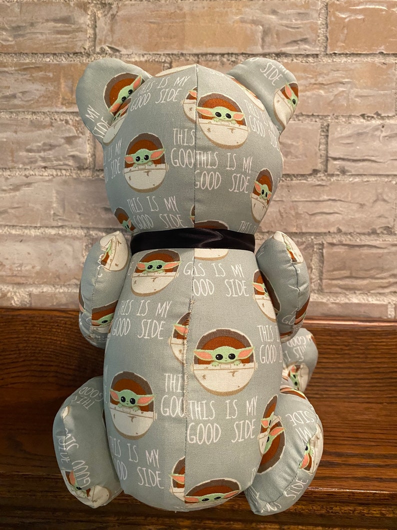K&ouml;nnte beinhalten: Ein Pl&uuml;schteddyb&auml;r mit einem hellblauen Stoff, der ein sich wiederholendes Muster eines gr&uuml;nen Babycharakters in einer Kapsel mit dem Text "This is my good side" auf hellblauem Hintergrund zeigt. Der B&auml;r hat ein schwarzes Band um den Hals.