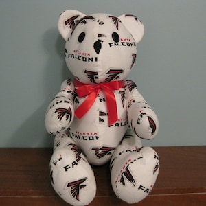 Puede incluir: Un oso de peluche blanco con una pajarita roja. El oso está cubierto de un patrón de logotipos de los Atlanta Falcons en negro y rojo. El oso está sentado sobre una superficie de madera.