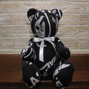 Könnte beinhalten: Ein schwarz-weißer Plüschteddybär mit einem weißen Band um den Hals. Der Bär trägt ein Stoffmuster mit dem Teamlogo und den Spielernamen der Las Vegas Raiders der NFL.