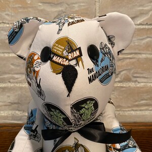 Op de afbeelding: Een witte teddybeer met zwarte knoopjesogen en een zwarte strik. De beer is bedekt met stof met een herhalend patroon van afbeeldingen uit de Star Wars-serie The Mandalorian. De stof bevat afbeeldingen van de Mandalorian, Grogu en de tekst "The Mandalorian" en "I've been looking for you".