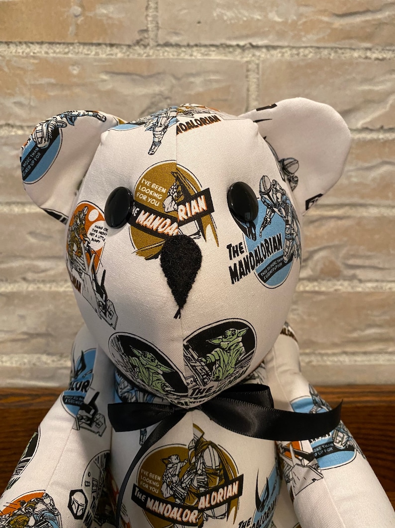 Op de afbeelding: Een witte teddybeer met zwarte knoopjesogen en een zwarte strik. De beer is bedekt met stof met een herhalend patroon van afbeeldingen uit de Star Wars-serie The Mandalorian. De stof bevat afbeeldingen van de Mandalorian, Grogu en de tekst "The Mandalorian" en "I've been looking for you".