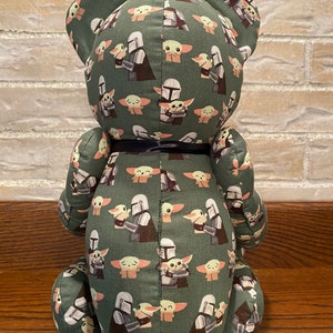 Puede incluir: Oso de peluche verde con un patr&oacute;n repetido de un personaje mandaloriano sosteniendo un beb&eacute; Yoda. El oso tiene una cinta negra alrededor del cuello.