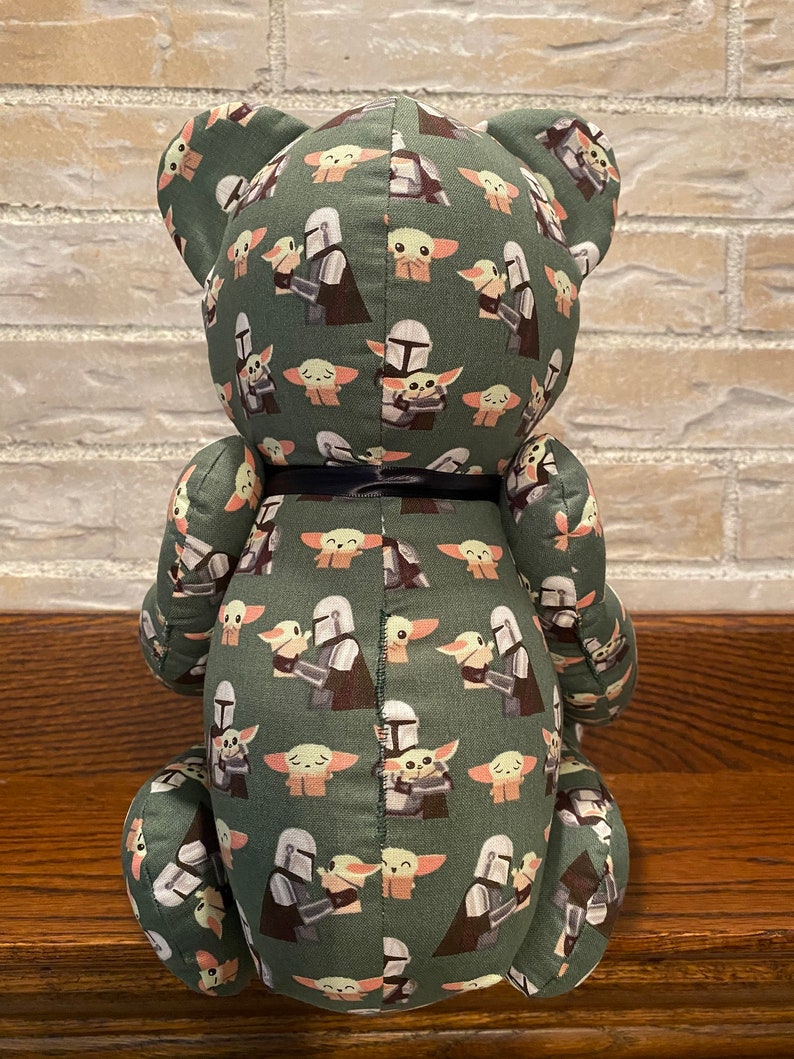 Puede incluir: Oso de peluche verde con un patr&oacute;n repetido de un personaje mandaloriano sosteniendo un beb&eacute; Yoda. El oso tiene una cinta negra alrededor del cuello.