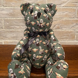 Puede incluir: Un oso de peluche verde con una cinta negra alrededor del cuello. El oso est&aacute; cubierto de un patr&oacute;n de peque&ntilde;as im&aacute;genes de un personaje alien&iacute;gena verde y un guerrero mandaloriano.