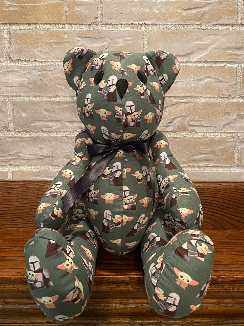 Puede incluir: Un oso de peluche verde con una cinta negra alrededor del cuello. El oso est&aacute; cubierto de un patr&oacute;n de peque&ntilde;as im&aacute;genes de un personaje alien&iacute;gena verde y un guerrero mandaloriano.