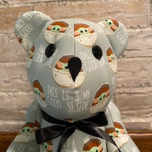 K&ouml;nnte beinhalten: Ein Pl&uuml;schteddyb&auml;r mit einem hellblauen Stoff, der ein sich wiederholendes Muster eines gr&uuml;nen Charakters in einer braunen Kapsel mit dem Text "This is my good side" zeigt. Der B&auml;r hat schwarze Knopfaugen und eine schwarze Schleife.