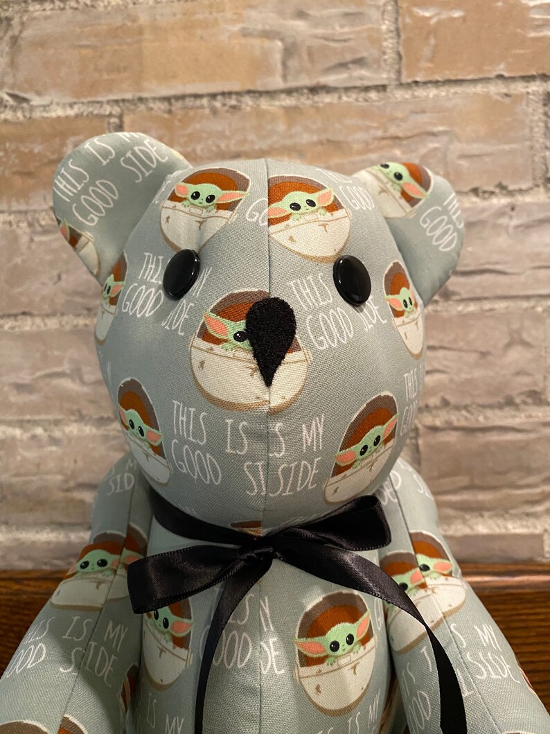 K&ouml;nnte beinhalten: Ein Pl&uuml;schteddyb&auml;r mit einem hellblauen Stoff, der ein sich wiederholendes Muster eines gr&uuml;nen Charakters in einer braunen Kapsel mit dem Text "This is my good side" zeigt. Der B&auml;r hat schwarze Knopfaugen und eine schwarze Schleife.