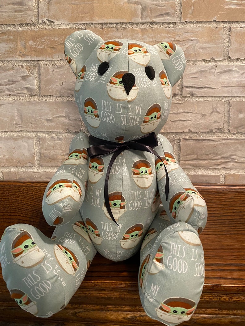 K&ouml;nnte beinhalten: Ein grauer Pl&uuml;schteddyb&auml;r mit einer schwarzen Schleife. Der B&auml;r ist mit einem Muster eines gr&uuml;nen Alien-Charakters bedeckt, auf dem der Text "This is my good side" wiederholt auf dem Stoff steht.