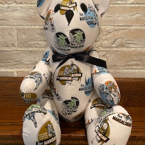 Op de afbeelding: Een witte teddybeer met een zwarte strik. De beer is bedekt met een stoffen print van verschillende sc&egrave;nes uit de Star Wars-serie The Mandalorian. De stoffen print bevat de tekst "The Mandalorian" en "The Mandalorian" in verschillende lettertypen en maten.