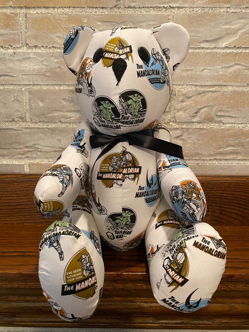 Op de afbeelding: Een witte teddybeer met een zwarte strik. De beer is bedekt met een stoffen print van verschillende sc&egrave;nes uit de Star Wars-serie The Mandalorian. De stoffen print bevat de tekst "The Mandalorian" en "The Mandalorian" in verschillende lettertypen en maten.