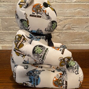 Op de afbeelding: Een witte pluche teddybeer met een zwarte neus en ogen. De beer is bedekt met een stoffen print met de woorden "The Mandalorian" en afbeeldingen van personages uit de Star Wars-serie.