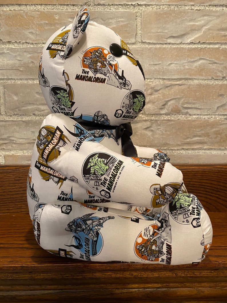 Op de afbeelding: Een witte pluche teddybeer met een zwarte neus en ogen. De beer is bedekt met een stoffen print met de woorden "The Mandalorian" en afbeeldingen van personages uit de Star Wars-serie.
