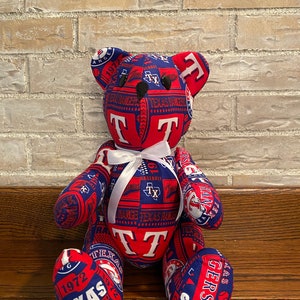 May include: A plush teddy bear made from fabric with a Texas Rangers theme. The bear is red and blue with the team's logo and other details. The bear is sitting on a wooden surface.