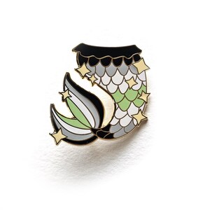 Puede incluir: Un pin de esmalte con borde dorado en forma de cola de sirena con un patrón de escamas en tonos negro, gris, blanco y verde claro. El diseño presenta varias estrellas doradas pequeñas de cuatro puntas alrededor de la cola y aletas.