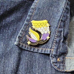 Puede incluir: Un pin de esmalte de color dorado con forma de cola de sirena. La cola está dividida en tres secciones: amarilla, blanca y morada, que representan los colores de la bandera del orgullo no binario. El pin tiene un acabado brillante y presenta pequeñas estrellas.