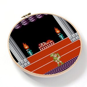 May include: A cross-stitch embroidery hoop with a pixelated image of a video game character standing on a platform with a red background. The character is green and has a sword. The background is black with a few orange flames.