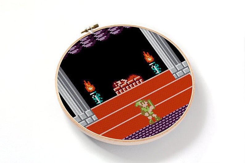 May include: A cross-stitch embroidery hoop with a pixelated image of a video game character standing on a platform with a red background. The character is green and has a sword. The background is black with a few orange flames.