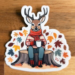 May include: A sticker featuring a deer wearing a red scarf and sweater, holding a white mug, and sitting on a tree stump. The deer is surrounded by colorful autumn leaves. The sticker has a white border.