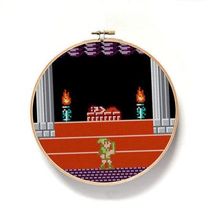 May include: A round, wooden embroidery hoop with a cross-stitch design of a video game scene. The scene features a green-clad character standing on a red platform with a brick structure behind them. There are two torches on either side of the brick structure.