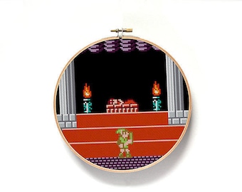 May include: A round, wooden embroidery hoop with a cross-stitch design of a video game scene. The scene features a green-clad character standing on a red platform with a brick structure behind them. There are two torches on either side of the brick structure.