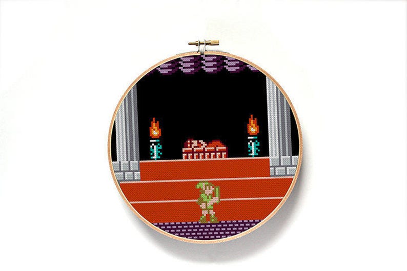 May include: A round, wooden embroidery hoop with a cross-stitch design of a video game scene. The scene features a green-clad character standing on a red platform with a brick structure behind them. There are two torches on either side of the brick structure.