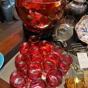May include: A set of 14 red glass cups and a large red glass punch bowl with a black base. The cups are arranged in a circle on a silver tray.