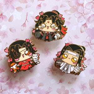May include: Three enamel pins depicting anime-style characters with black hair and gold detailing. Each pin features a different outfit and is decorated with pink hearts. One pin includes a black cat, another a white cat, and the third a bunny.
