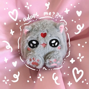 Puede incluir: Un juguete de peluche gris en forma de gato con ojos grandes y un corazón rojo en el pecho. El juguete está rodeado de corazones blancos y el texto "¿adoptarme?" sobre un fondo rosa.