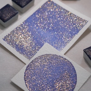 May include: Two samples of iridescent purple watercolour paint with glitter. One sample is a square shape and the other is a circle.