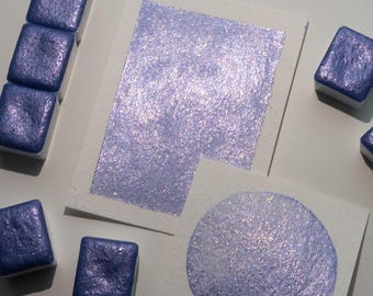 May include: A set of six watercolor paint pans in a deep blue color with a shimmery finish. The paint is shown on two white paper squares, one with a square swatch and the other with a round swatch.