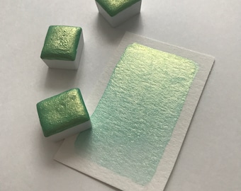May include: Three small square pans of green metallic watercolor paint. The paint is a light green with a shimmery finish. A sample of the paint is shown on a white paper.