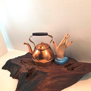 May include: A polished copper tea kettle with a black handle and a matching lid, positioned next to a beige ceramic hand sculpture featuring red painted fingernails and a blue base, all resting on a dark, textured piece of natural wood.
