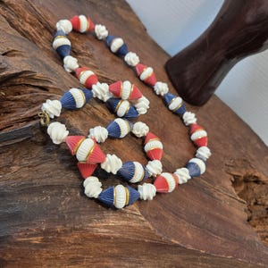 May include: A beaded necklace featuring alternating red, white, and navy blue beads. The beads are shaped like ribbed cones and spheres, separated by small gold-toned metal rings. The necklace includes a simple metal clasp closure at the end.