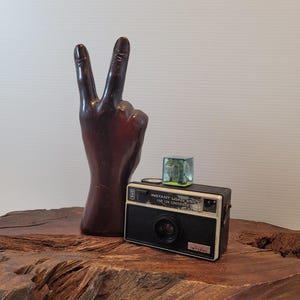 May include: A vintage black and white Imperial Instant Load 800 camera with a flash attachment. A dark brown hand sculpture is in the background, with the index and middle fingers raised in a peace sign. The items are on a wooden surface.