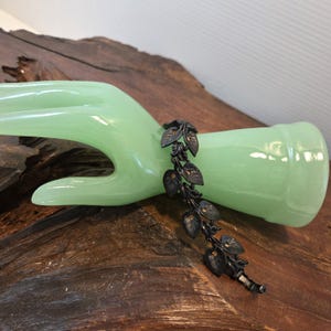 May include: A dark, matte black metal bracelet featuring a series of interconnected leaf-shaped links. Each individual leaf link is detailed with etched veins and a small, centered amber-colored stone, creating a continuous botanical design.