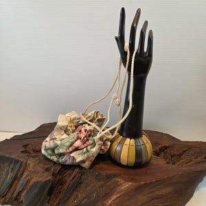 May include: A small floral drawstring bag with a cream-colored cord, resting on a wooden surface. The bag features a pattern of pink, green, and cream-colored flowers. A black hand-shaped display stand holds a cream-colored necklace.