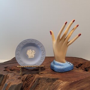 May include: A light blue decorative plate with a raised relief design, displayed on a small gold-colored stand. Next to it is a hand-shaped jewelry holder with red painted fingernails, resting on a blue base. The items are on a wooden surface.