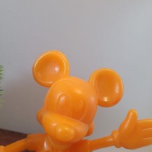 May include: A bright orange plastic figurine of Mickey Mouse. Mickey is wearing a short-sleeved shirt and has his right arm outstretched.