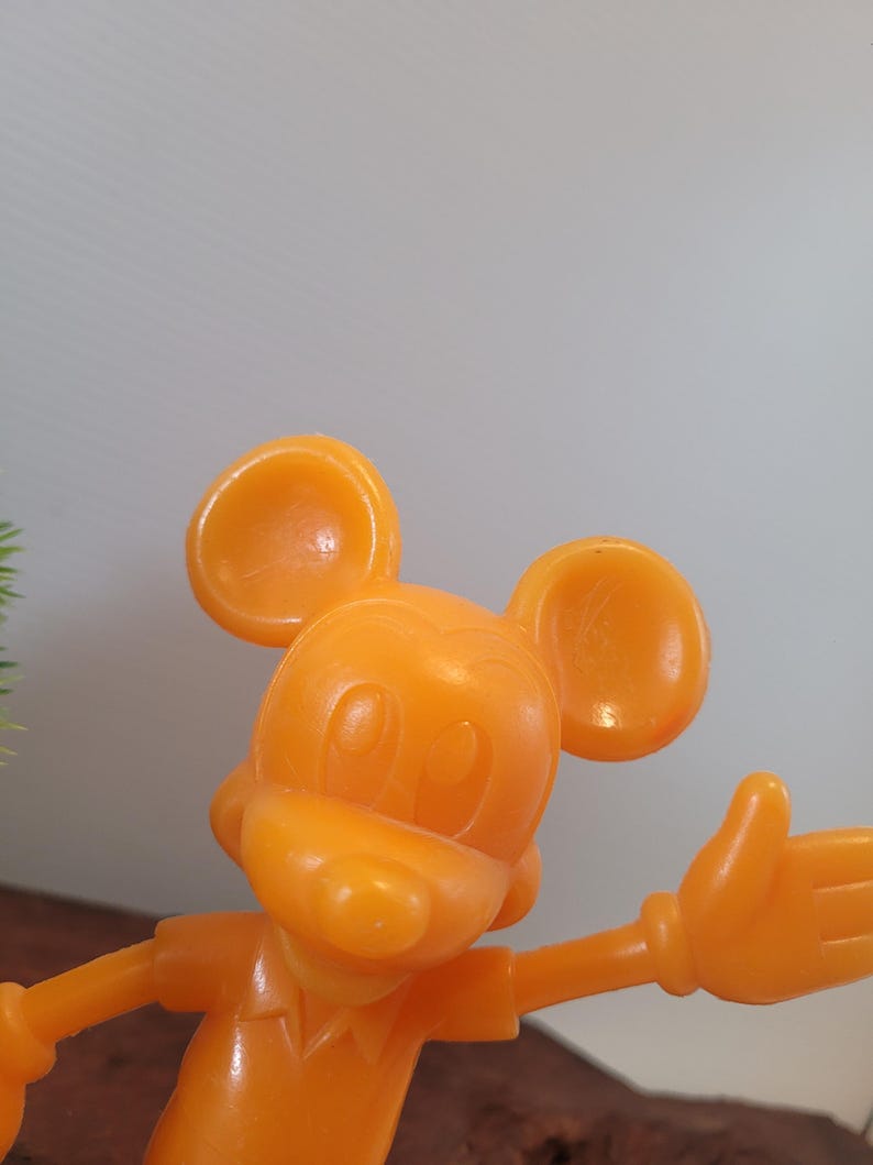 May include: A bright orange plastic figurine of Mickey Mouse. Mickey is wearing a short-sleeved shirt and has his right arm outstretched.