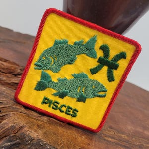 May include: A yellow square patch with a red border, featuring two green embroidered fish and the word "PISCES" in green embroidery.