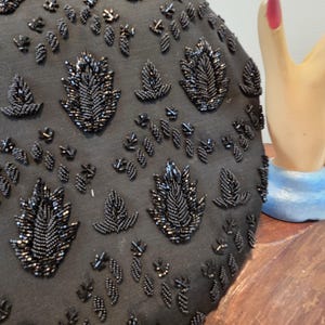 Pode incluir: Bolsa de noite preta com contas e intrincados desenhos florais e de folhas. A superf&iacute;cie da bolsa &eacute; coberta por pequenas contas escuras, criando um efeito texturizado e brilhante. O design apresenta folhas e formas florais com contas de v&aacute;rios tamanhos.