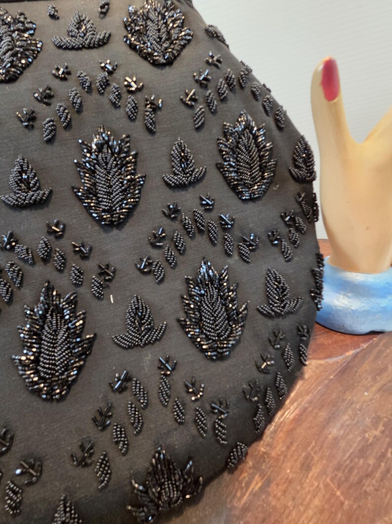 Pode incluir: Bolsa de noite preta com contas e intrincados desenhos florais e de folhas. A superf&iacute;cie da bolsa &eacute; coberta por pequenas contas escuras, criando um efeito texturizado e brilhante. O design apresenta folhas e formas florais com contas de v&aacute;rios tamanhos.